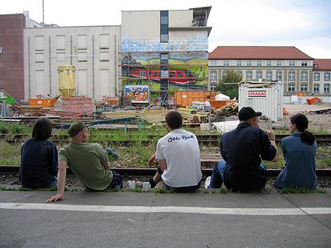 Teilnehmer des Projekts „Graffiti Workshop 2004“ sitzen am Bahnsteig vor den Gleisen und blicken auf die Wand des InterCityHotel Erfurt.