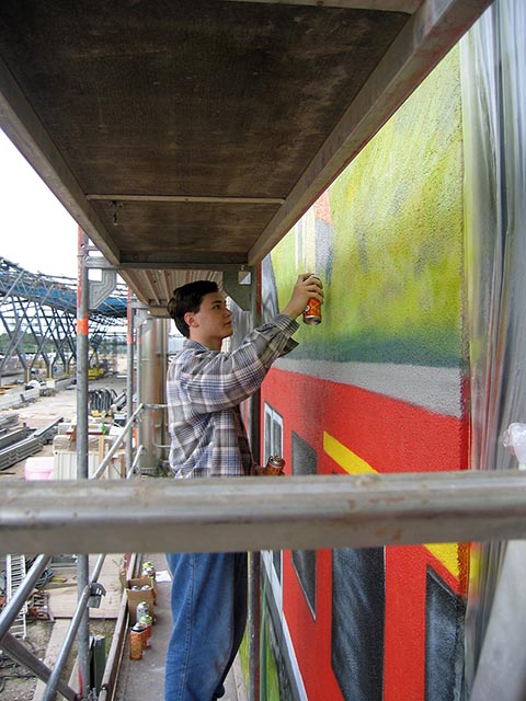 Alexander Babel sprüht mit der Spraydose Farbe auf und malt eine Familie – Vater mit Reisetasche, Mutter mit Regenschirm und Kind mit Hund, die gerade aus dem Zug ausgestiegen sind. „Graffiti Workshop 2004“.